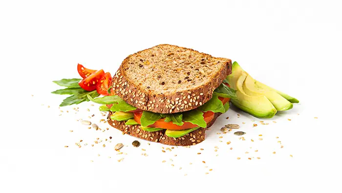 Un sandwich integral con aguacates, tomates, rúcula y semillas sobre un fondo blanco.
