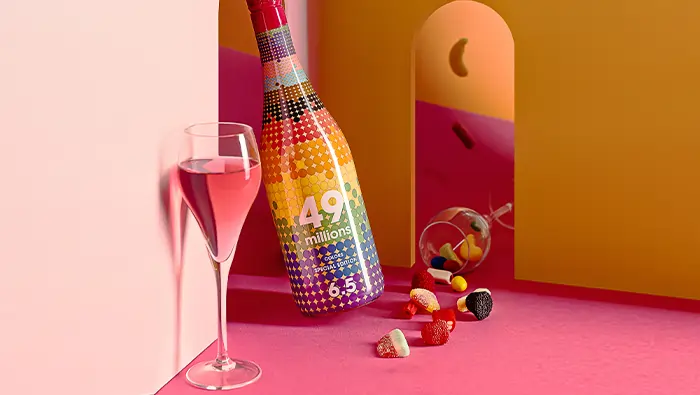 Una botella de vino y una copa en un entorno surrealista de colores rosa y amarillo con chuches coloridos.