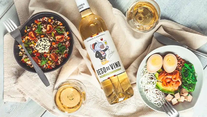 Botella de vino blanco y tazones de ensalada con tenedores sobre mantel individual con copas y cuchillos.