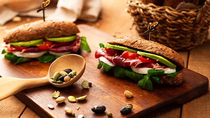 Dos sándwiches con jamón serrano, aguacate, tomate y espinacas sobre tabla de cortar con cuchara de madera.