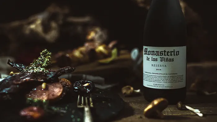 Una botella de vino junto a un plato de pizarra que contiene un guiso de conejo y ciruelas en un entorno oscuro con cepas de vid al fondo.