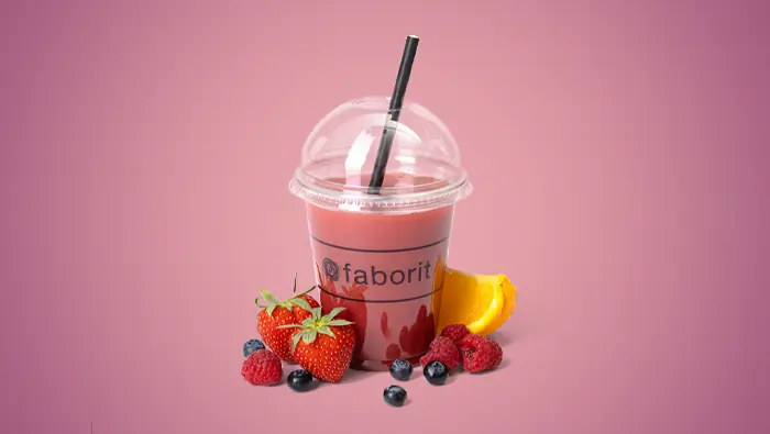 Un vaso de zumo rojo con pajita, un gajo de naranja, fresas, frambuesas y arandanos en fondo lila.