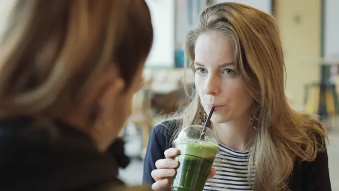 Una mujer tomando un batido verde en un café frente a otra mujer.
