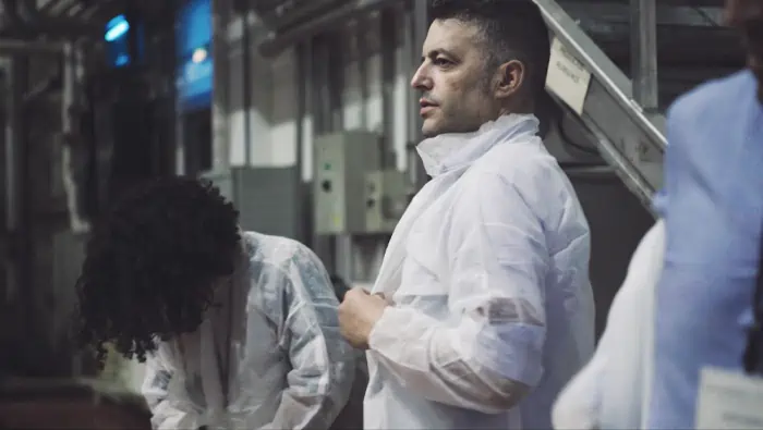 Un hombre en bata blanca junto a otras personas en un entorno industrial.