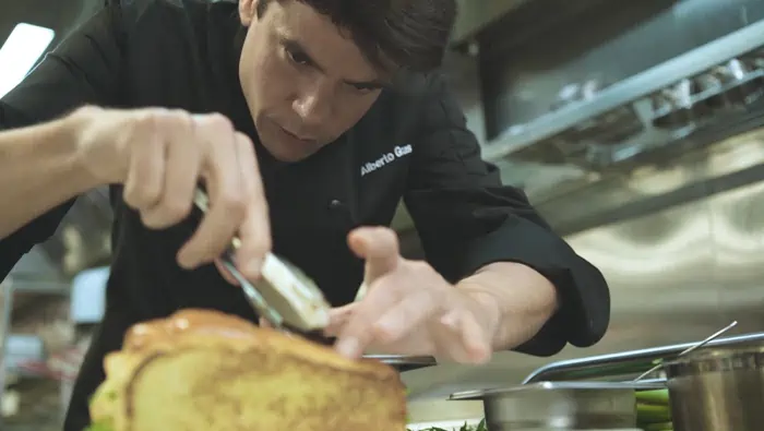 Un chef con camisa negra preparando un sandwich en una cocina profesional.